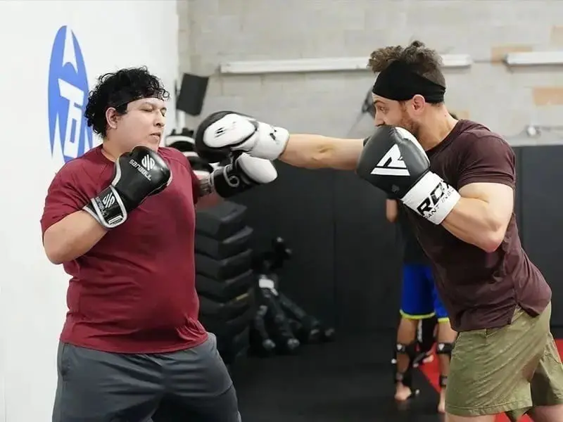 Two men practice controlled boxing combinations during a self-defense training session in a martial arts gym.