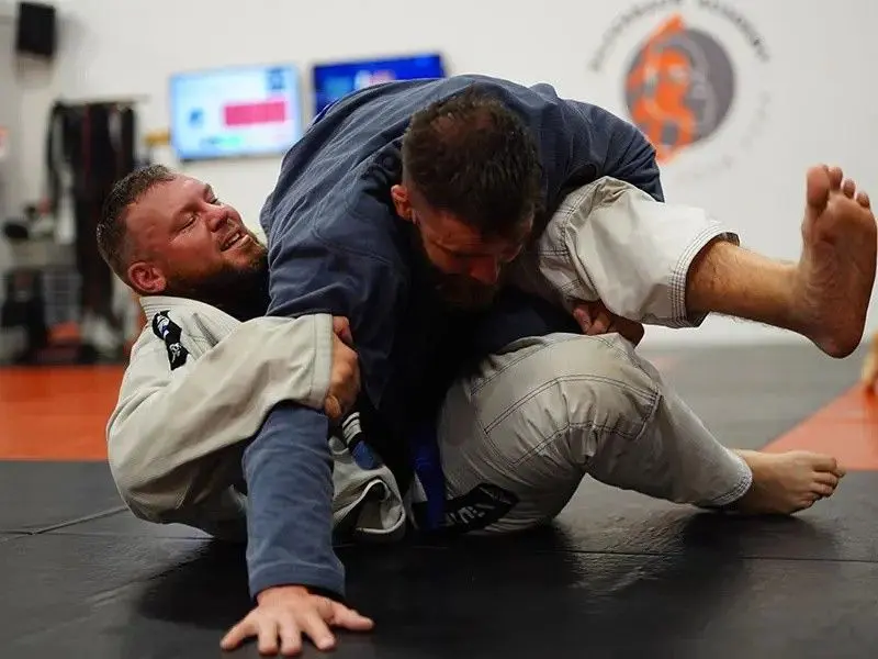 A dynamic action shot of two male athletes grappling on a gym mat during one of their self-defense classes, with one practitioner working for a sweep or reversal.